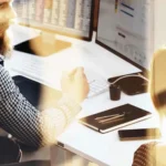 A bearded man in a checkered shirt sits at a desk with computer monitors, discussing b2b strategies with a woman whose back is turned to the camera. They are in a well-lit office of an SEO agency.