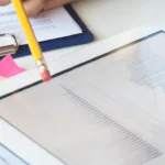 Two hands pointing with pencils at a digital tablet displaying colorful graphs and charts, possibly from a SaaS dashboard. Nearby are documents on a clipboard, additional colorful charts, and sticky notes, suggesting a business or financial meeting setting.